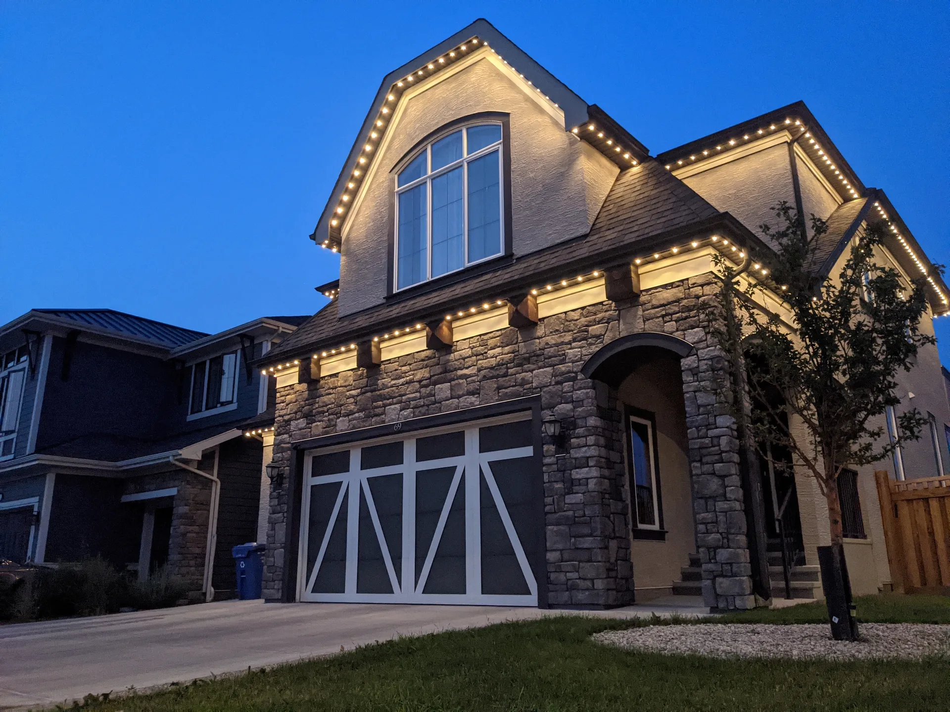Two-story stone and stucco home at dusk with warm white permanent LED lights tracing the rooflines, peaks, and garage