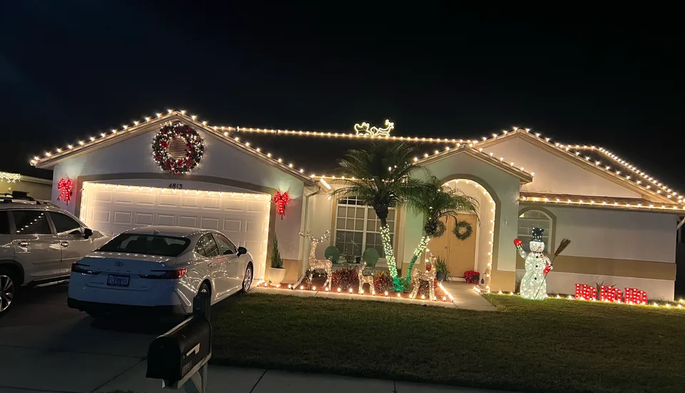 Florida home at night outlined in warm white C9 Christmas lights, with a lit wreath, red bows, palm trunks wrapped in lights, and glowing reindeer and a snowman on the lawn