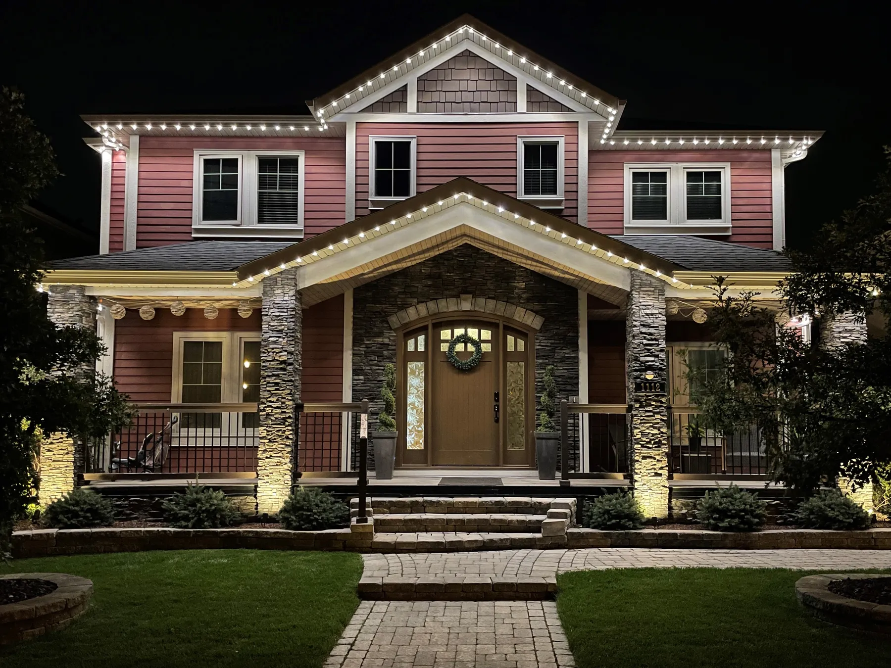 Craftsman-style home at night outlined in warm permanent roofline lighting with landscape uplighting on the stone columns and a lit front walkway