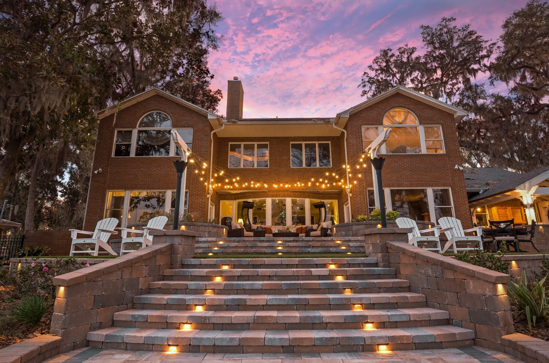 Dusk lakeside home with lit steps, string lights, pink sky, glowing windows