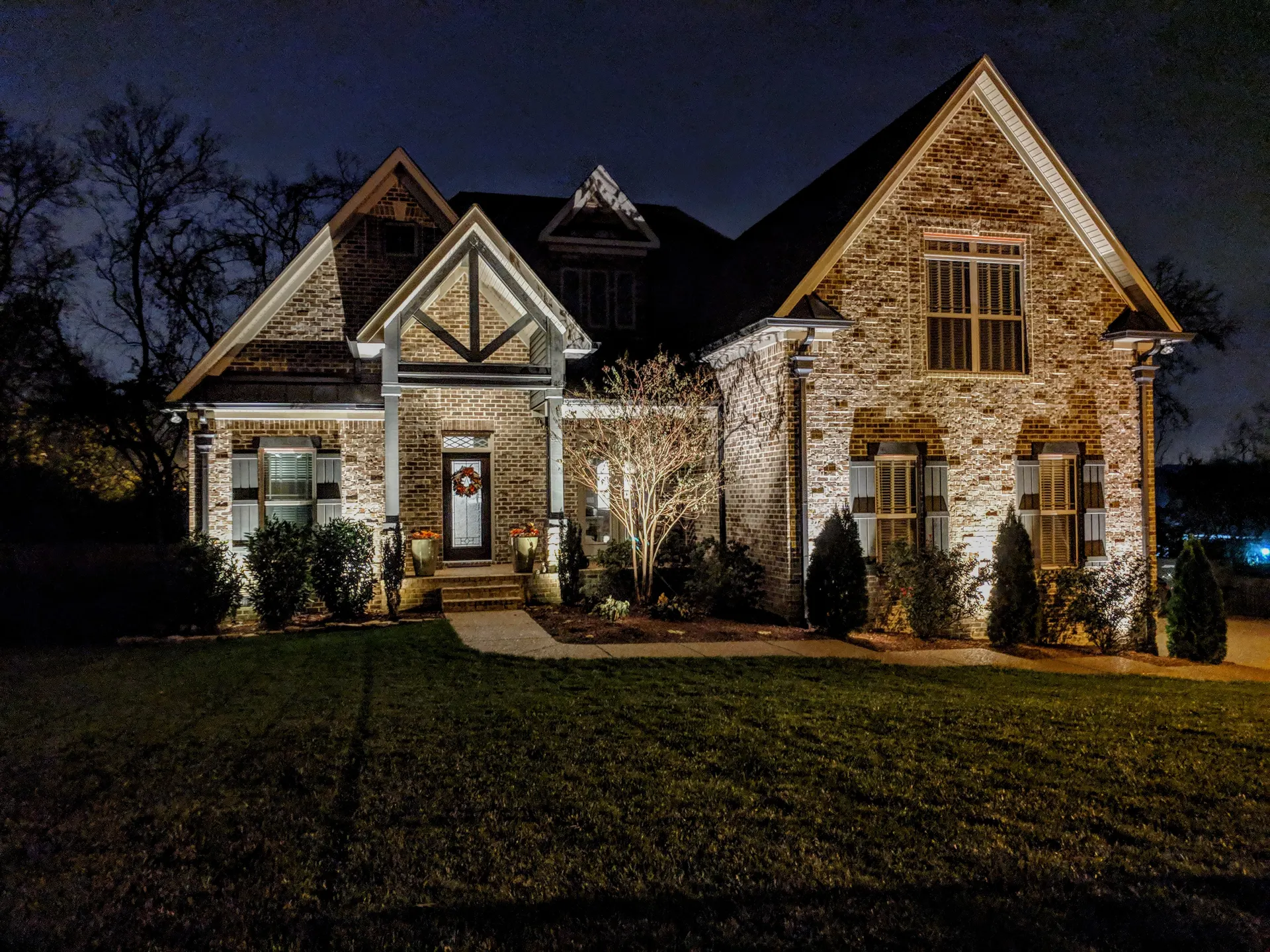 Night brick home with facade uplighting, lit windows, tree uplight