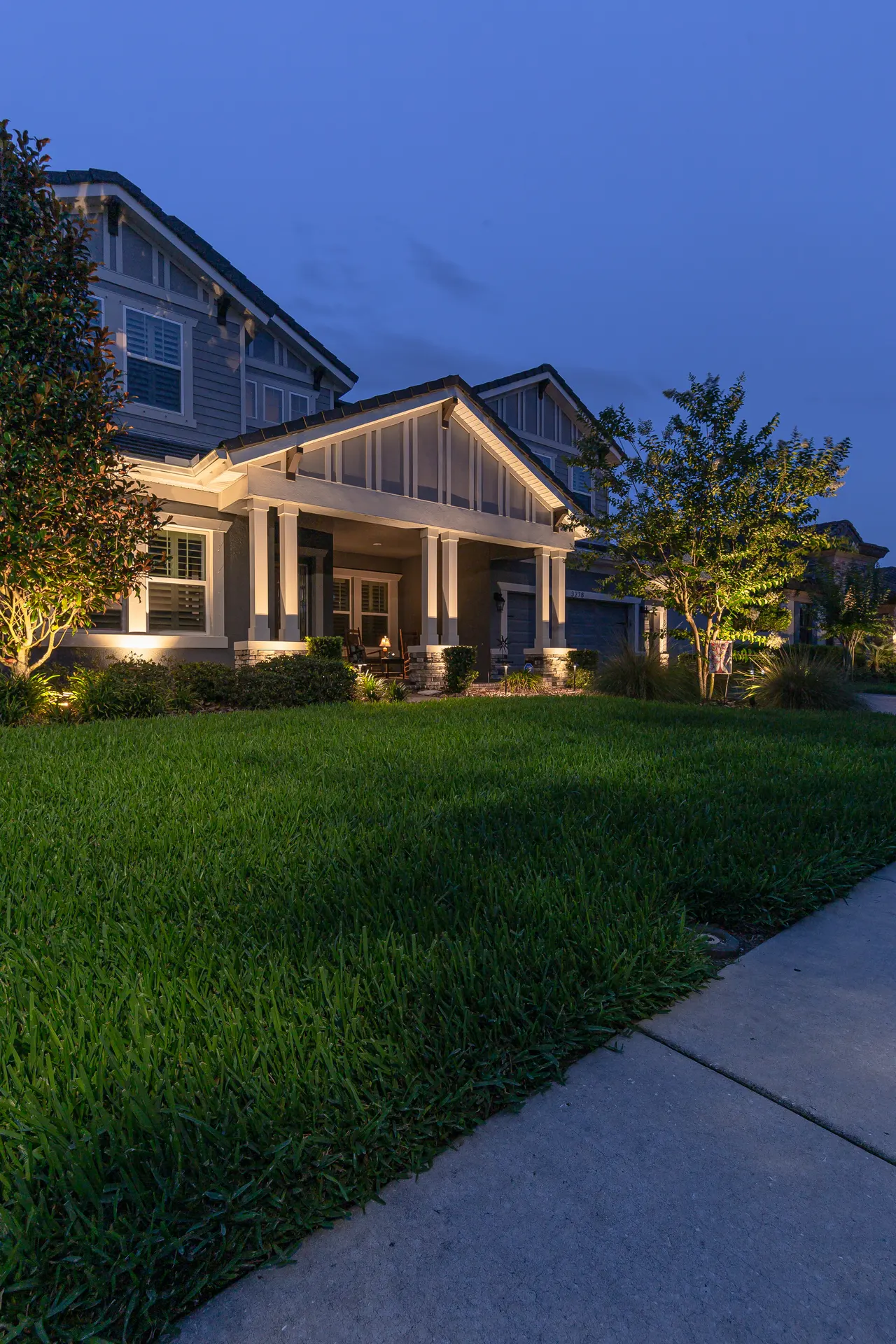 Craftsman home dusk, uplit trees and glowing porch, lush lawn, blue sky
