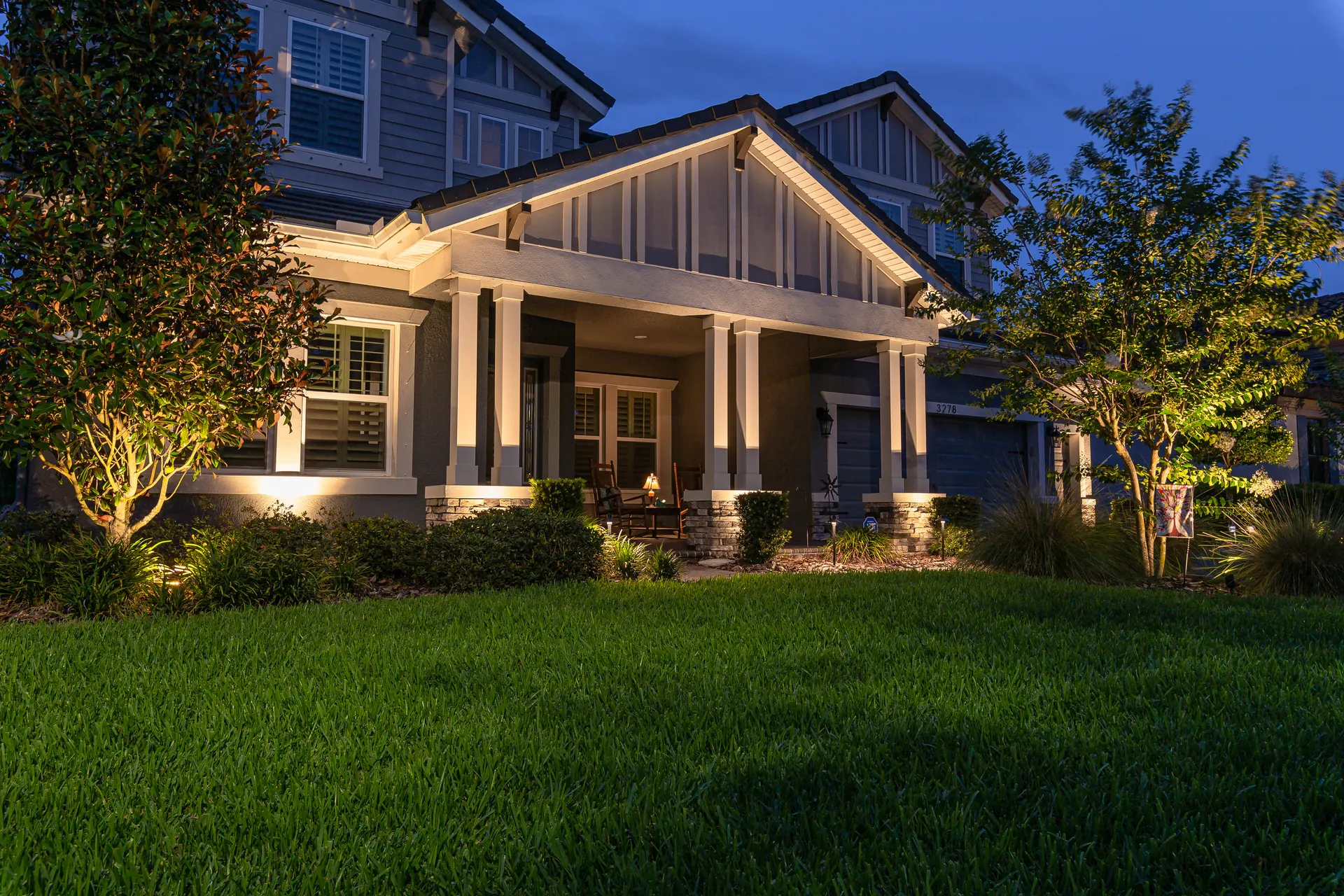 Craftsman porch with columns, warm uplighting on facade and tree, lush lawn