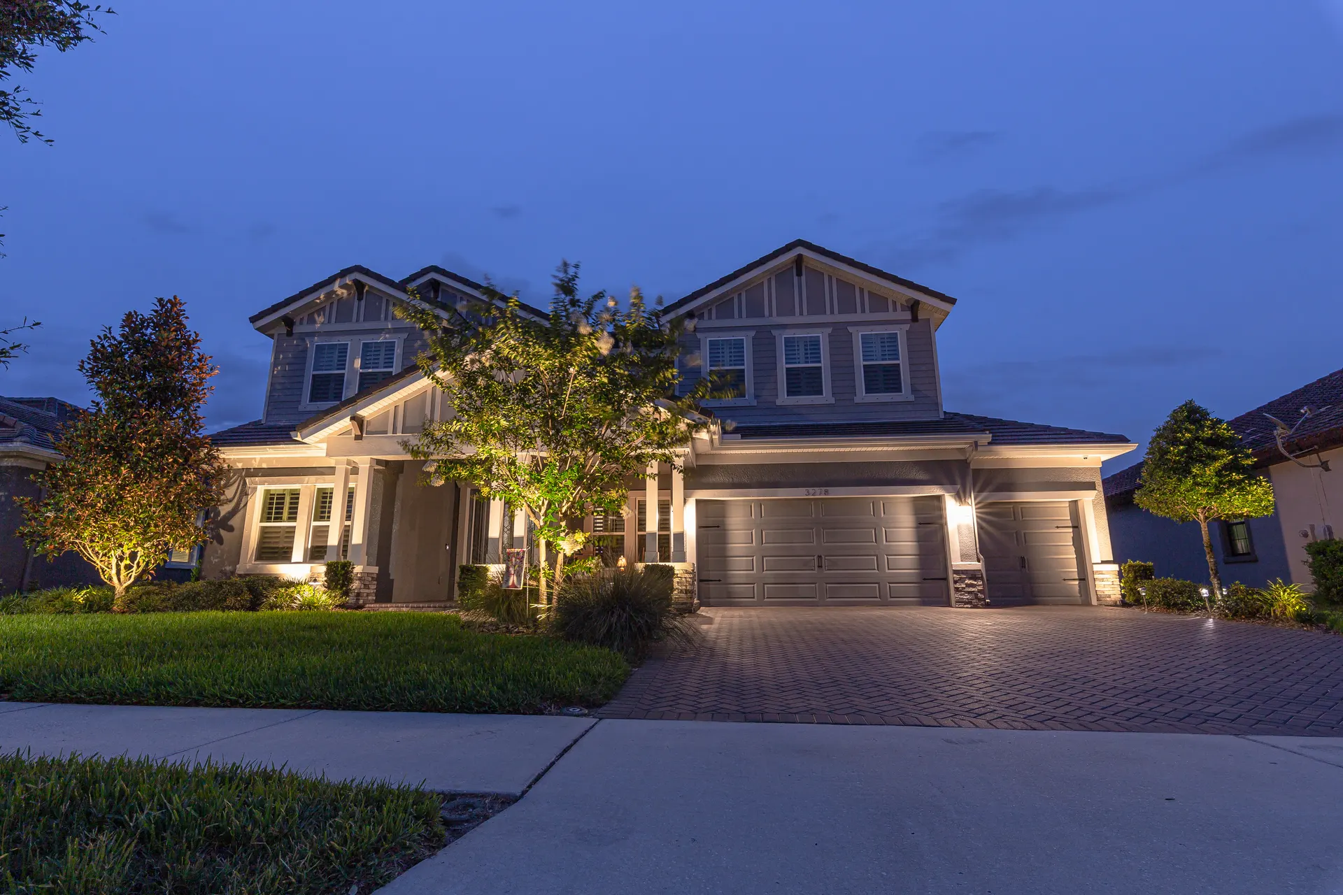 Two-story craftsman home at dusk, uplit trees, glowing entry, brick drive