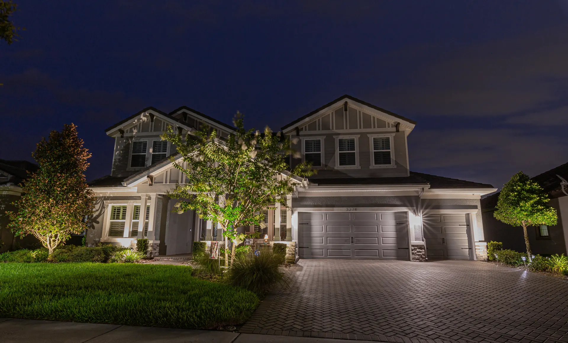Two-story home at night, uplit trees and facade, glowing garage, brick drive