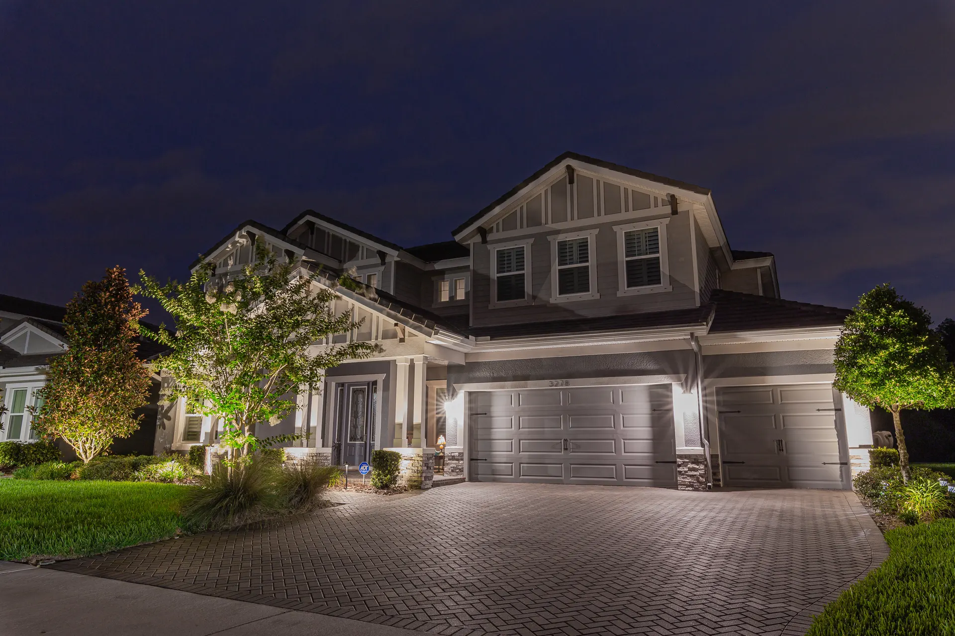 Gray home at dusk, uplit trees and entry, glowing garage, wide brick drive