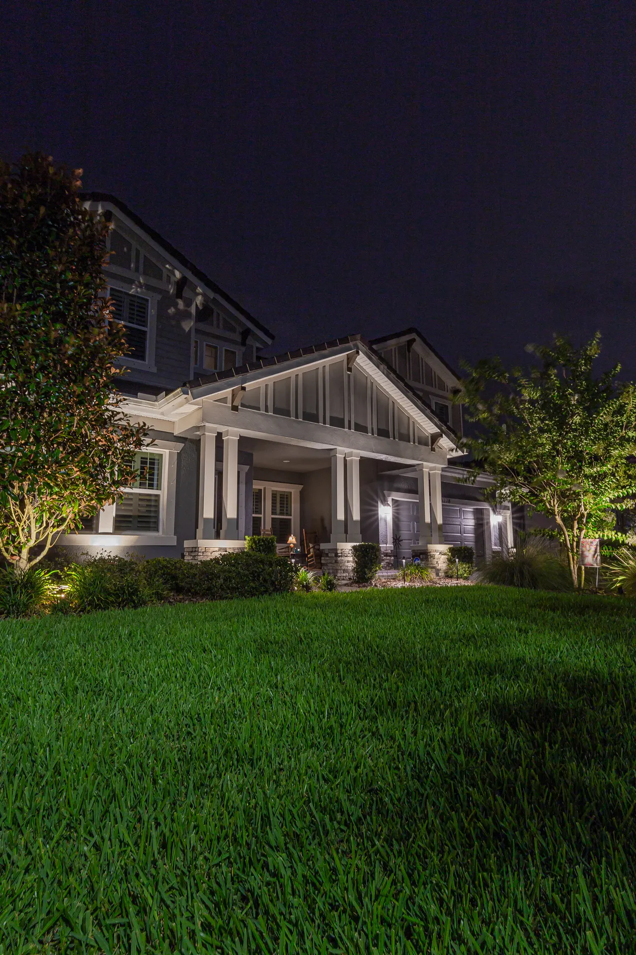Craftsman home night, uplit trees flanking lit porch, deep sky, lush lawn