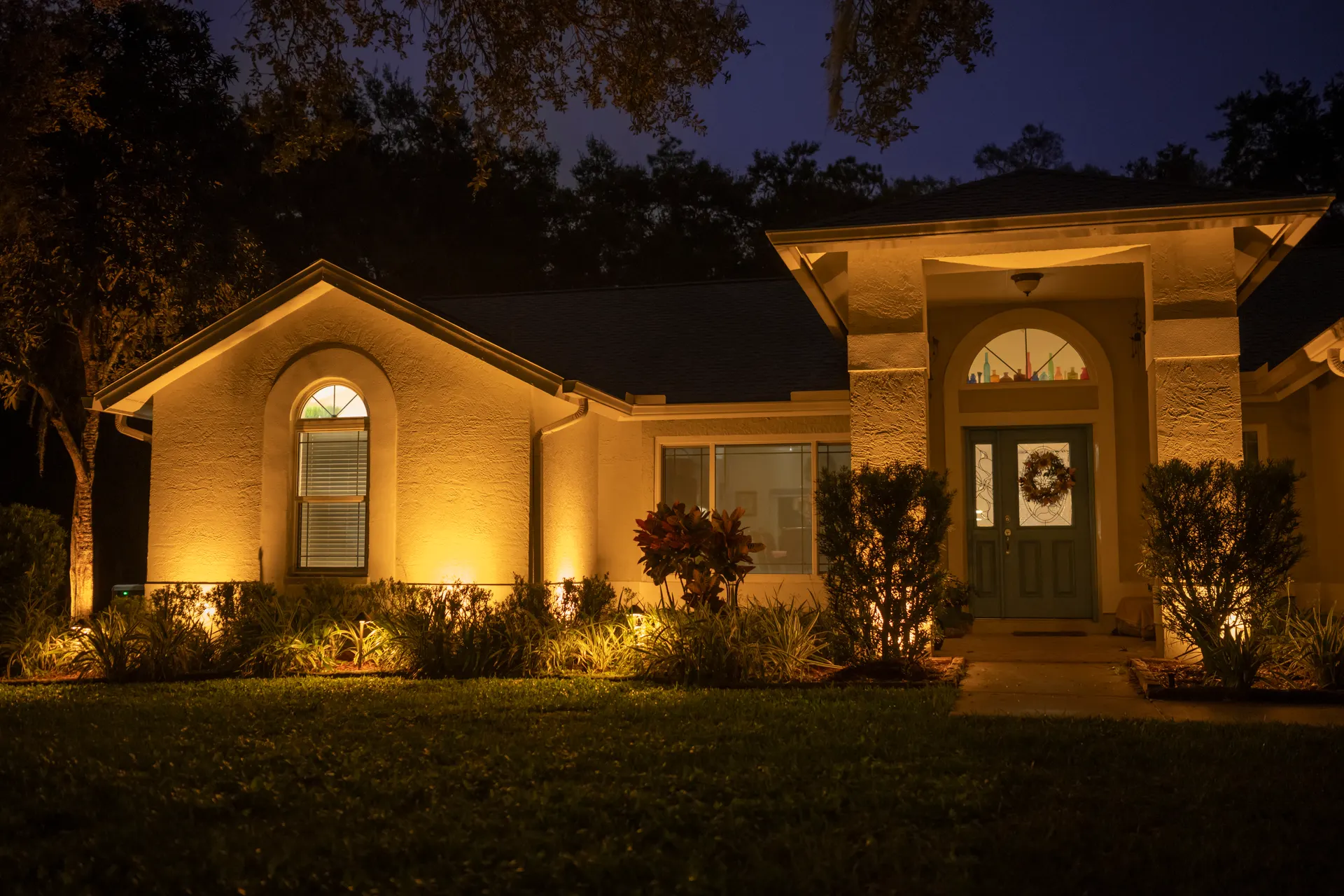 White brick ranch warm uplighting, glowing entry, twilight sky, lush beds