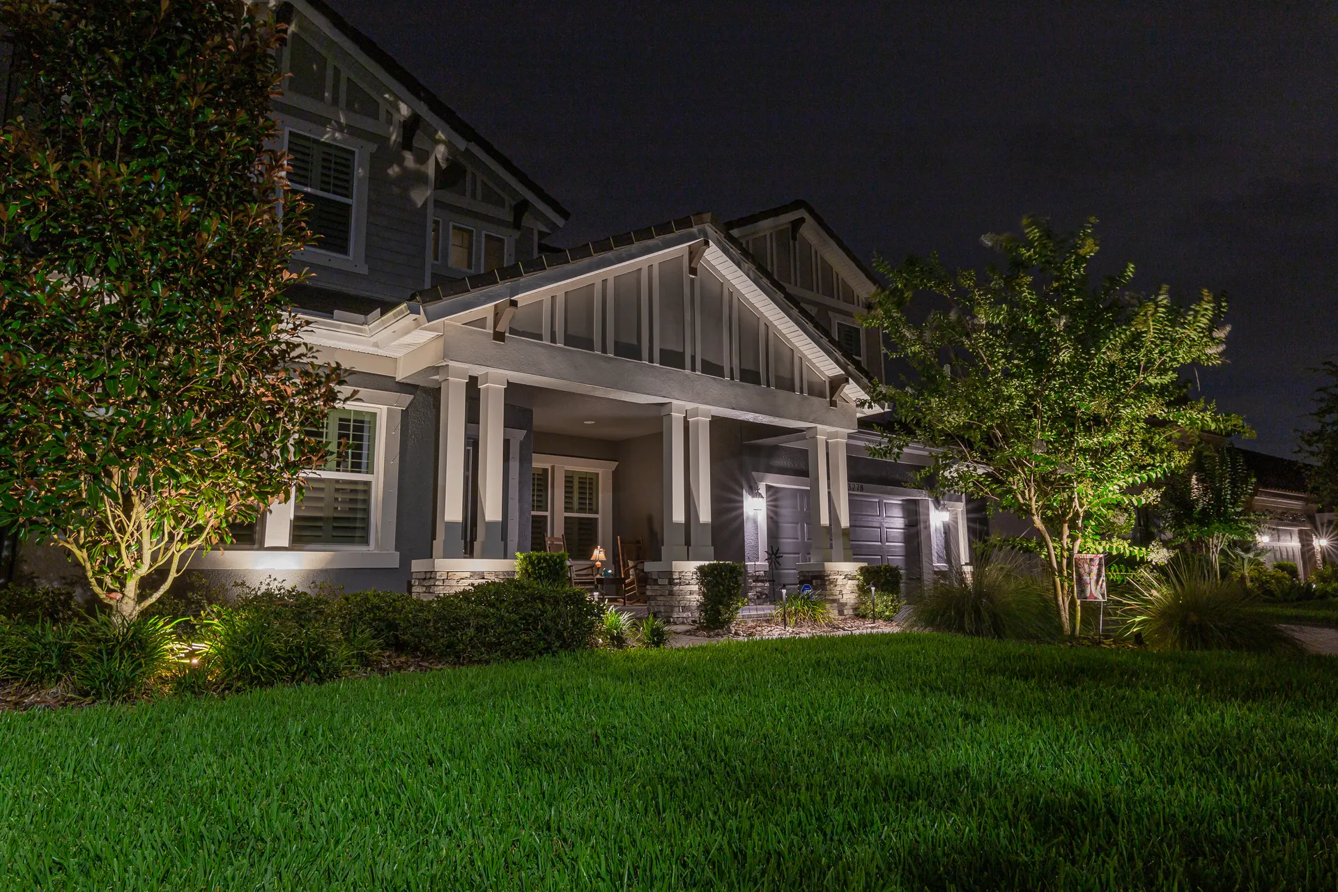 Craftsman porch at night, columns and trees uplit, lush lawn foreground