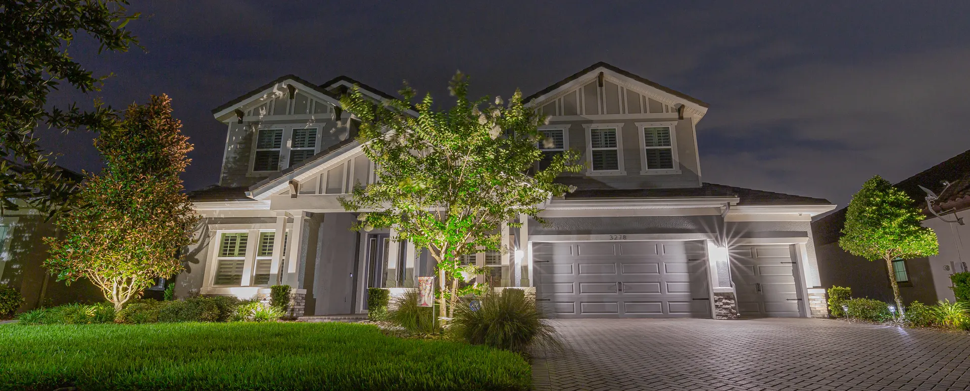 Two-story home at night, balanced facade wash, uplit trees, paver drive