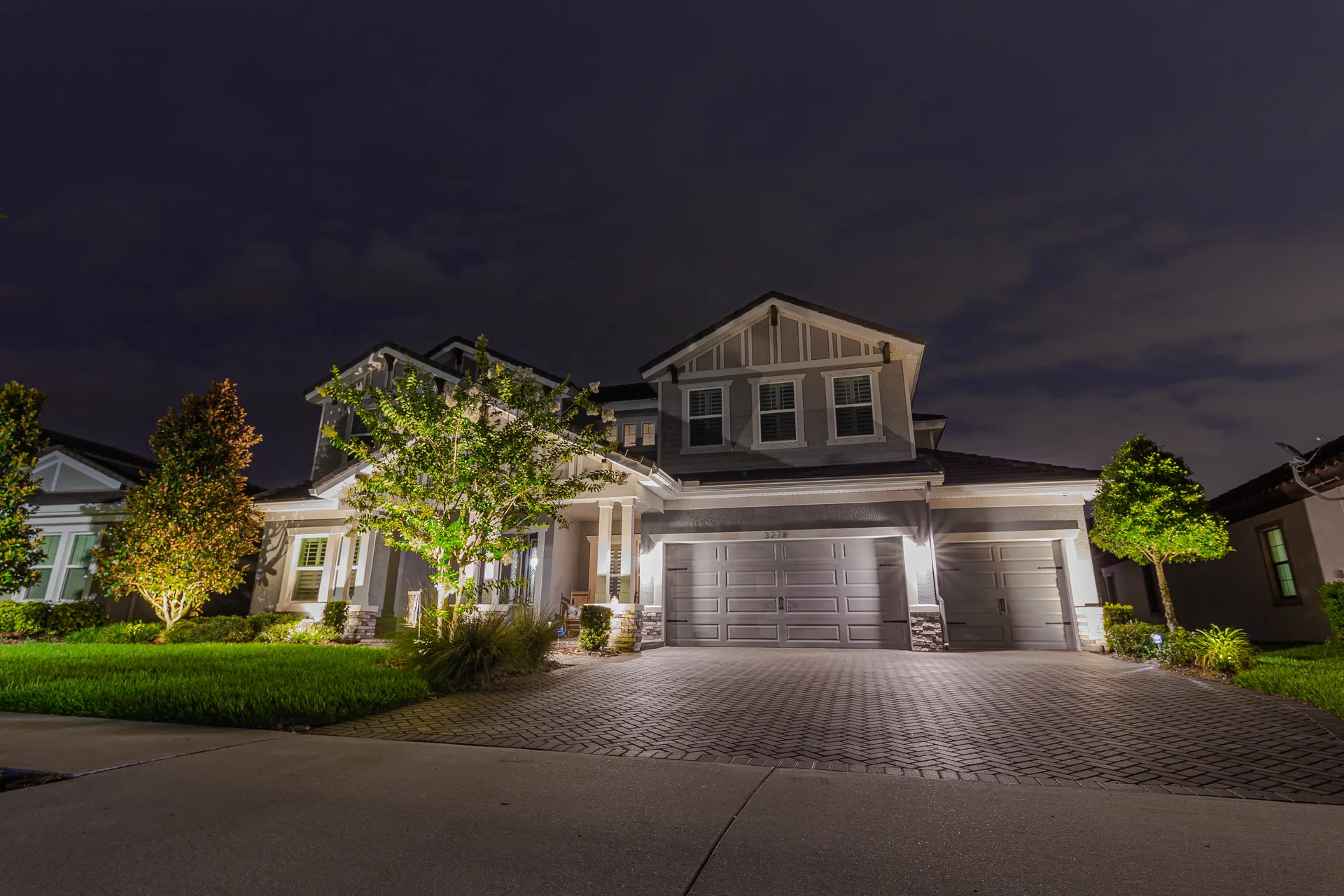 Craftsman two-story dusk, clean facade wash, uplit landscaping, paver driveway