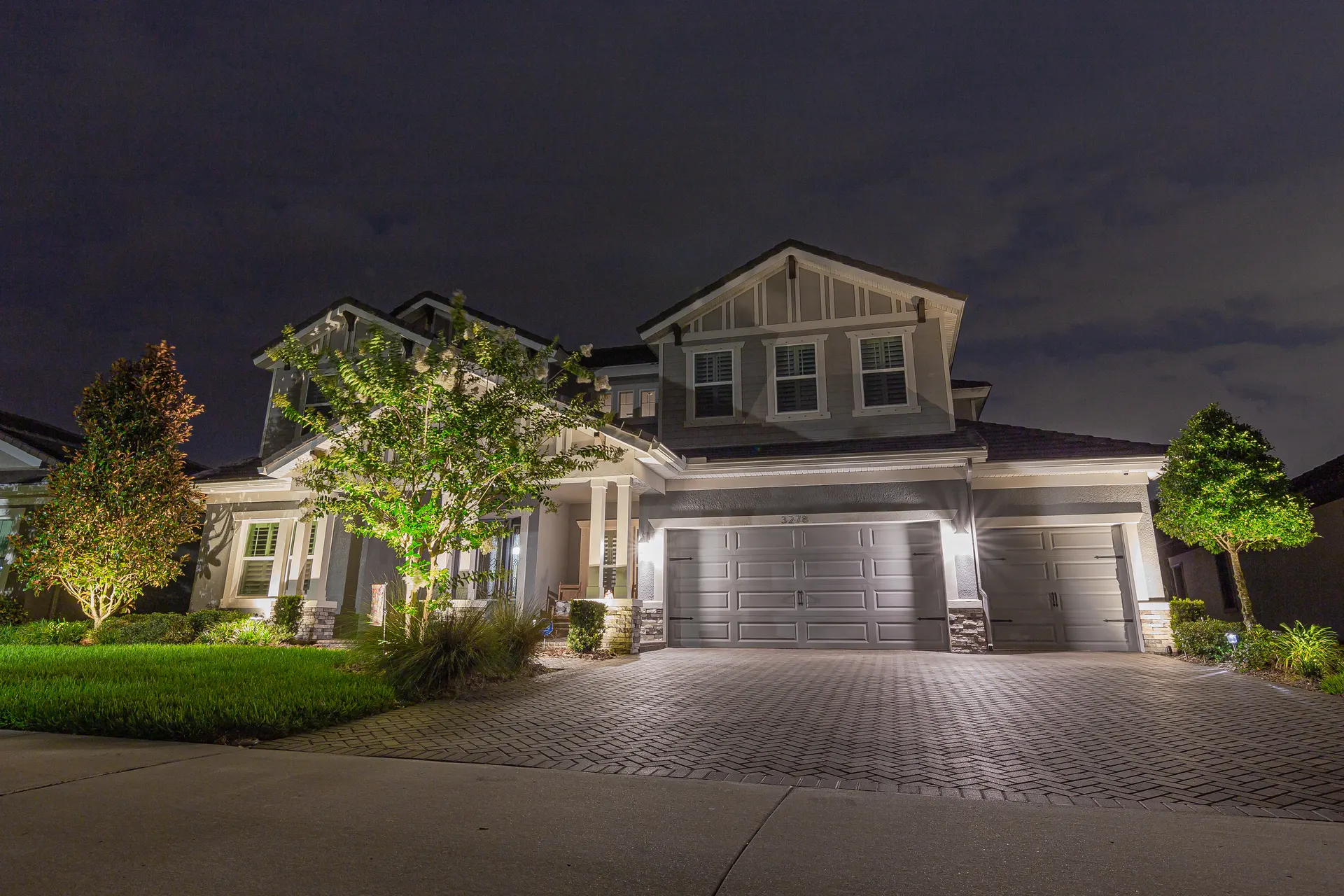 Two-story home at night, warm facade wash, uplit shrubs and trees