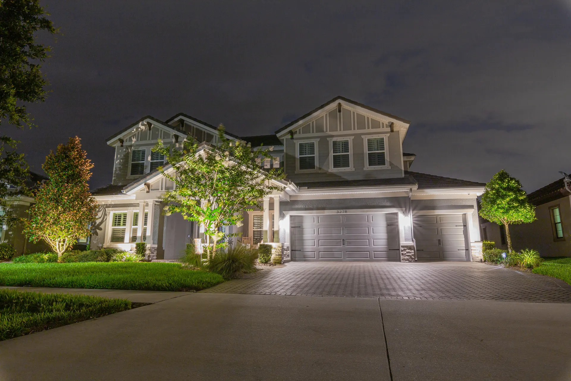 Wide two-story home night, balanced facade wash, uplit trees, paver drive