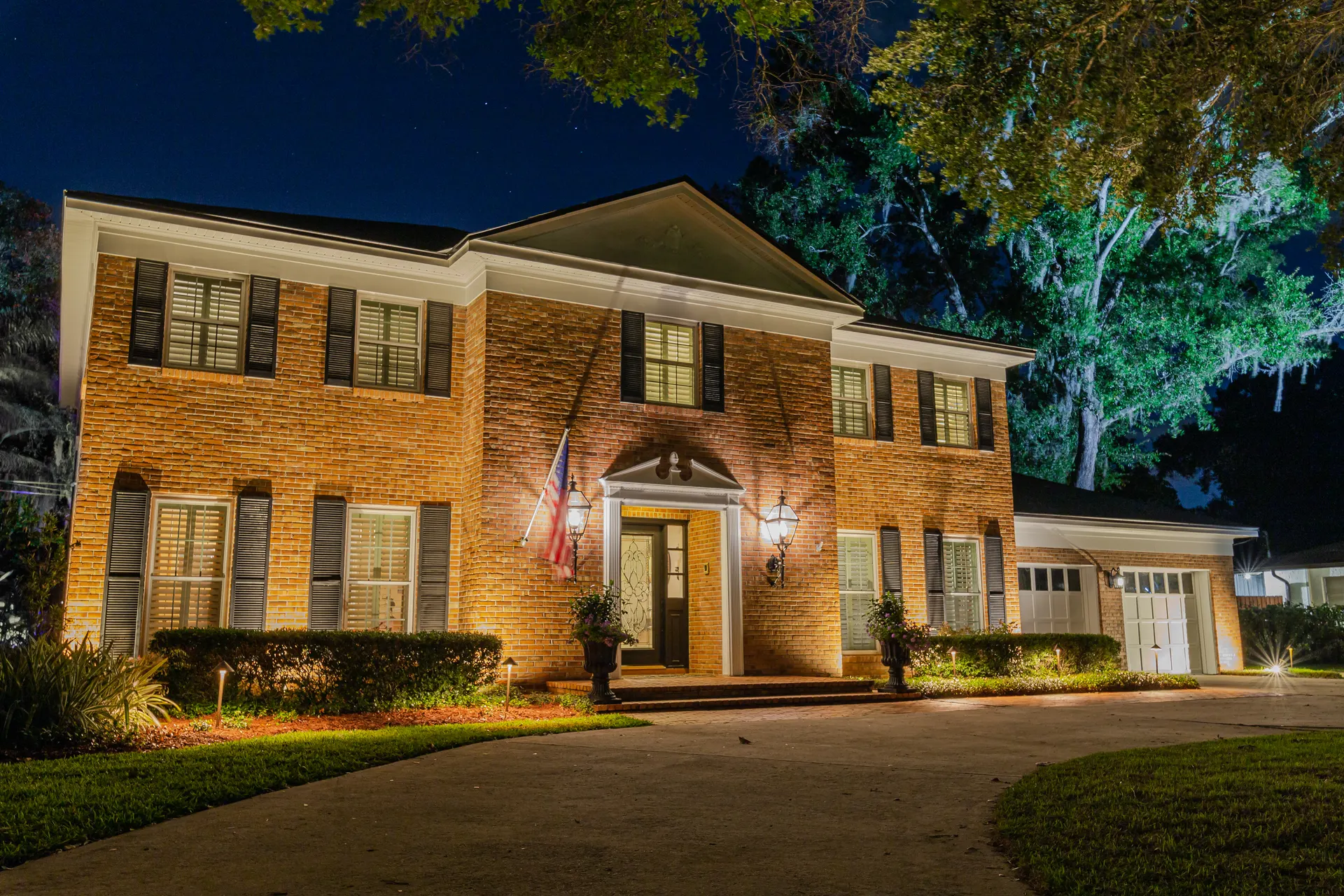 Colonial brick home night, warm facade wash, uplit landscaping, columns lit