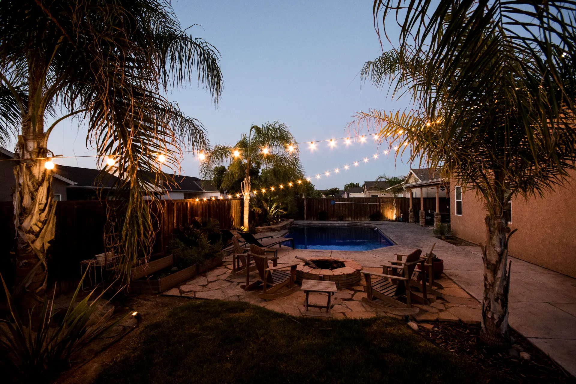 Dusk backyard pool, string lights, palms, fire pit seating area