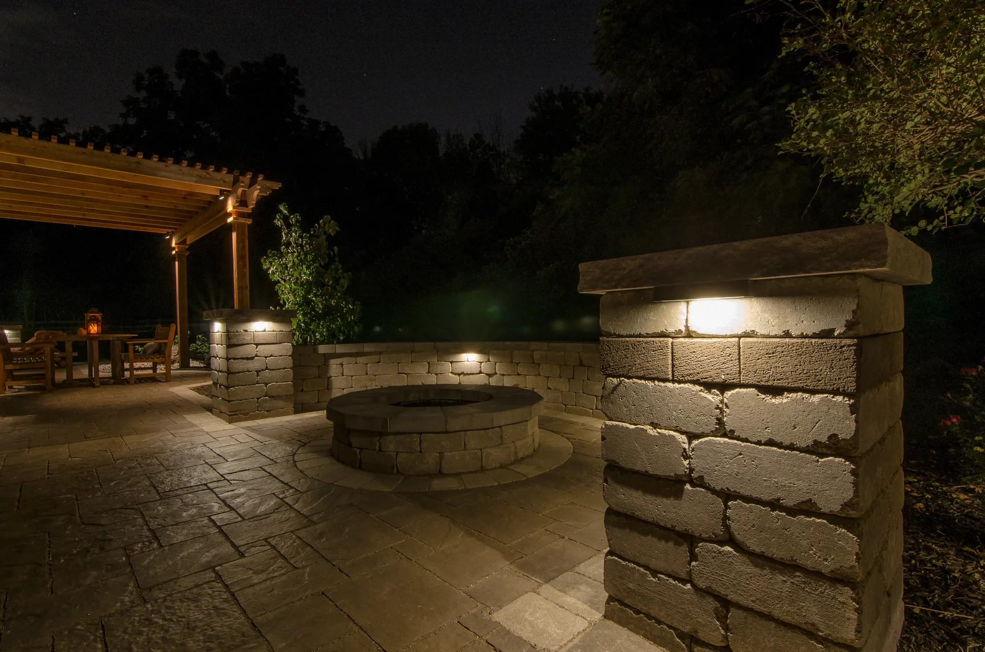 Paver patio with lit fire pit, pergola, glowing column-cap wall lights, night