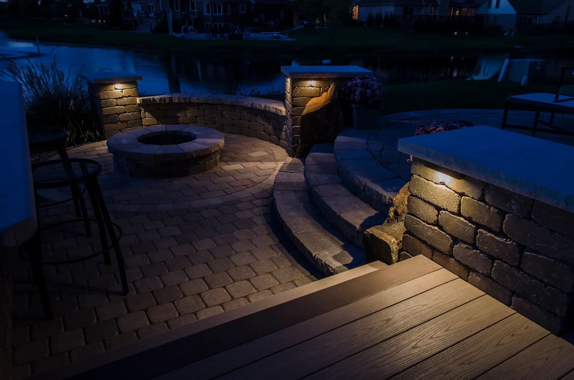 Lakeside paver patio with a fire pit, water feature, and wall and step lights at dusk