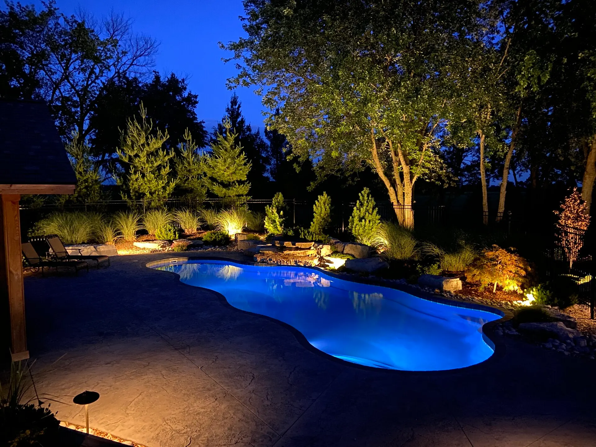 Dusk backyard, glowing blue pool, uplit trees, path-lit patio