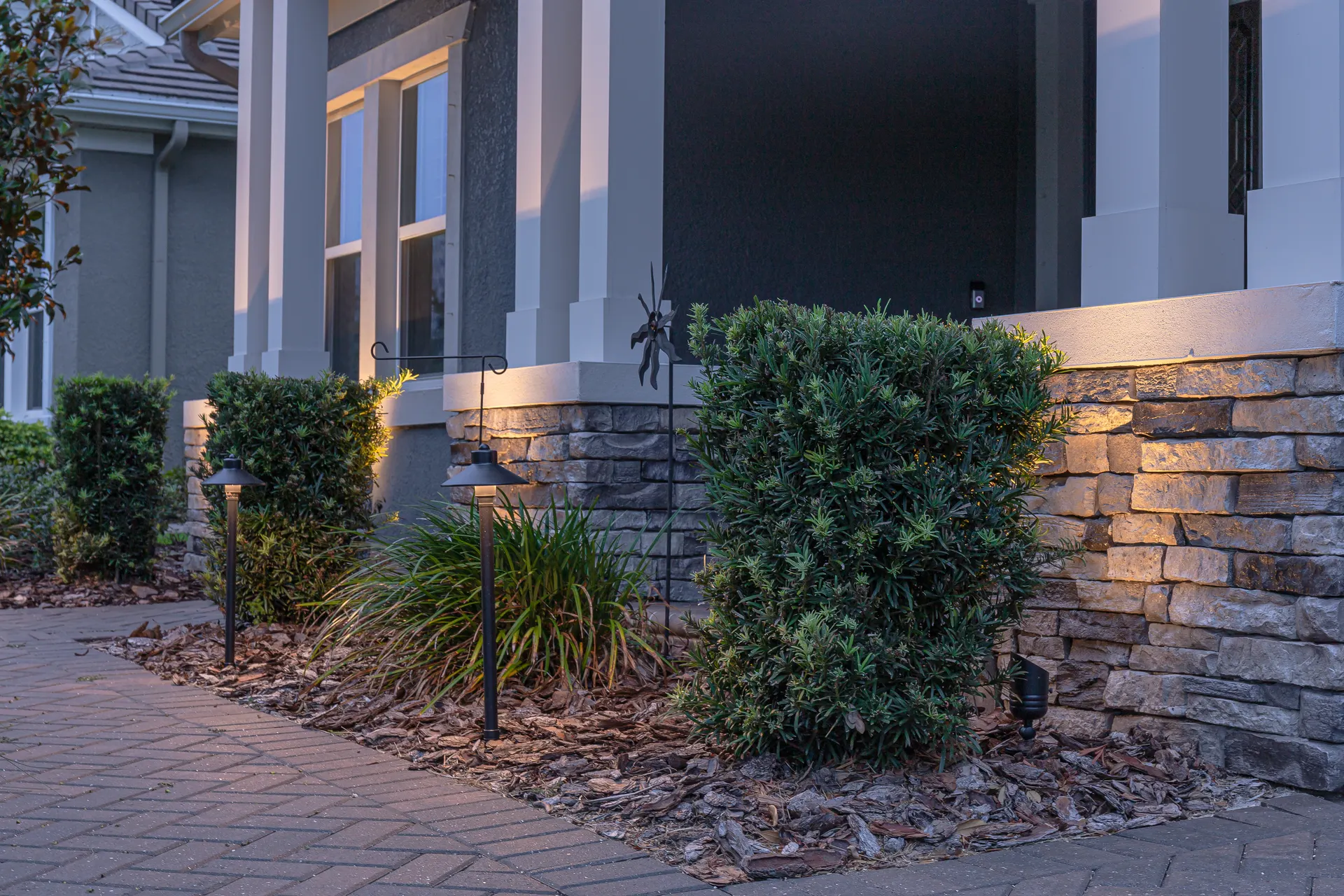 Stone-base porch with path lights and uplit shrubs, refined dusk entry detail