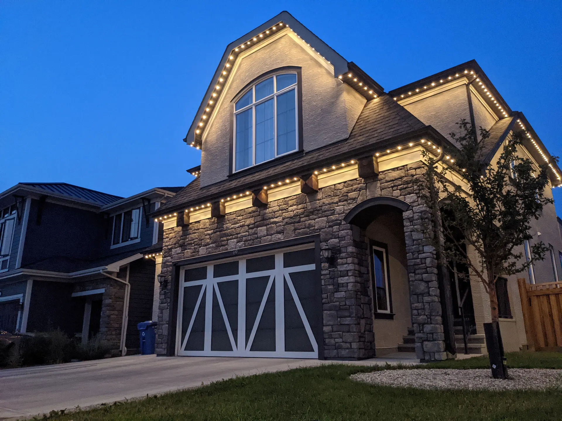 Stone two-story home at dusk with warm white permanent lighting outlining the gables, eaves, and garage