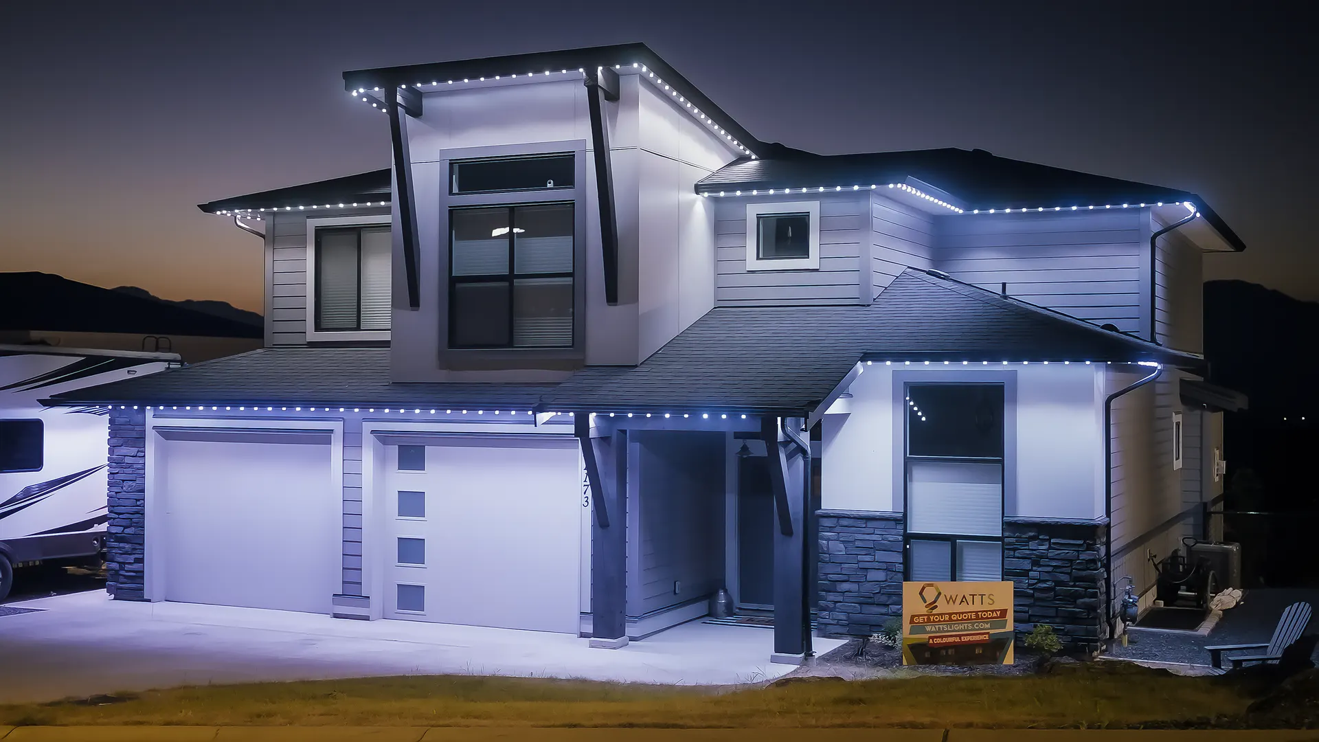 Modern home eaves and peaks outlined in cool white at dusk, small yard sign