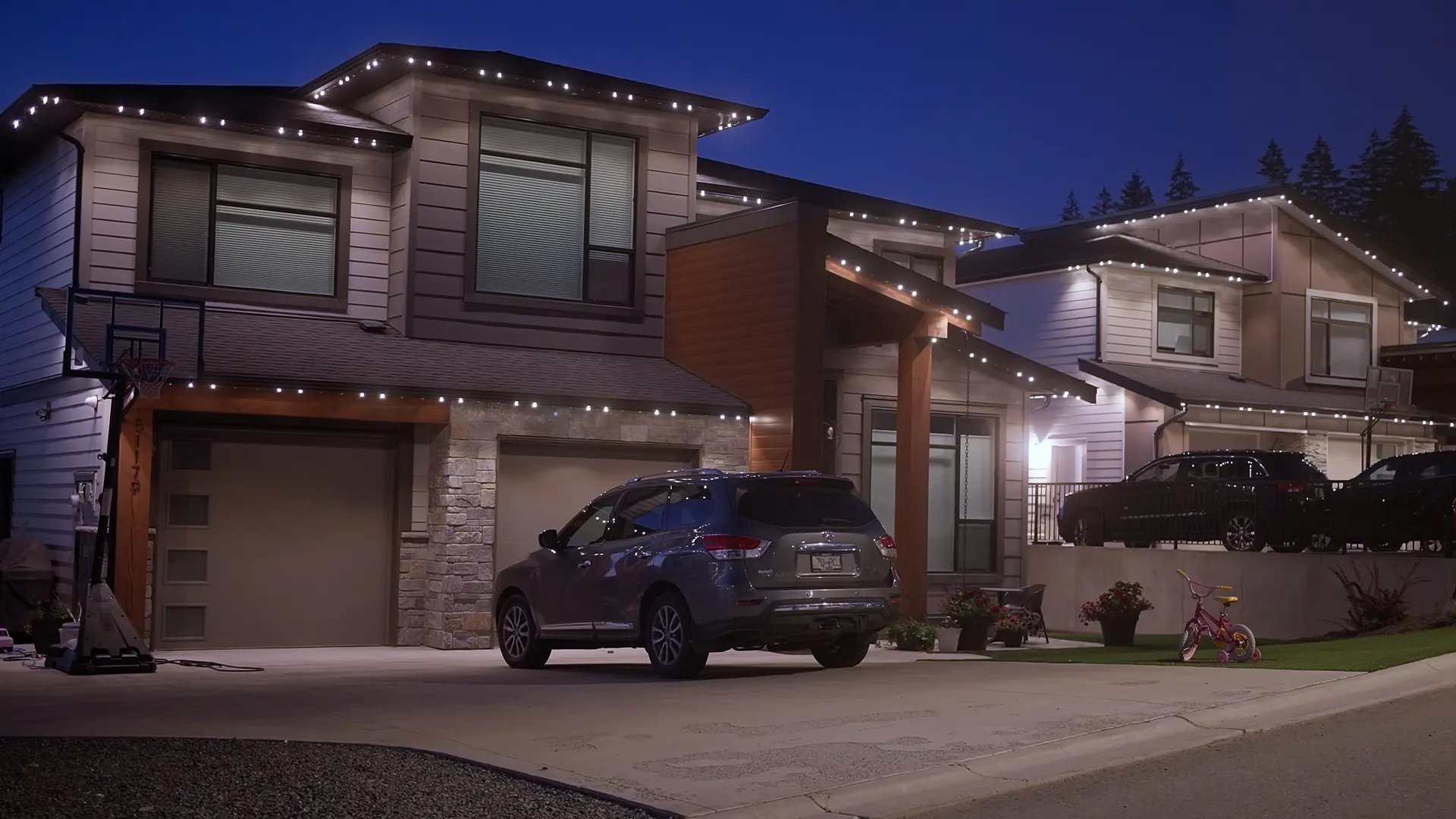 Hillside homes eaves outlined in warm white at dusk, SUV in driveway