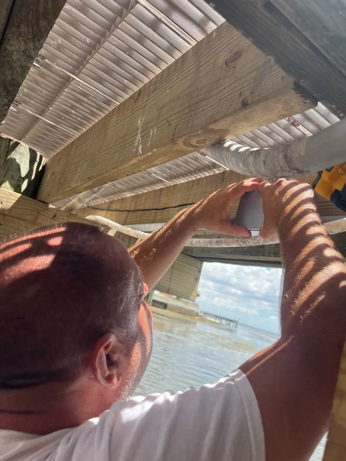 Installer mounting a small low-voltage light fixture to the underside of a dock frame, water and a neighboring dock visible beyond