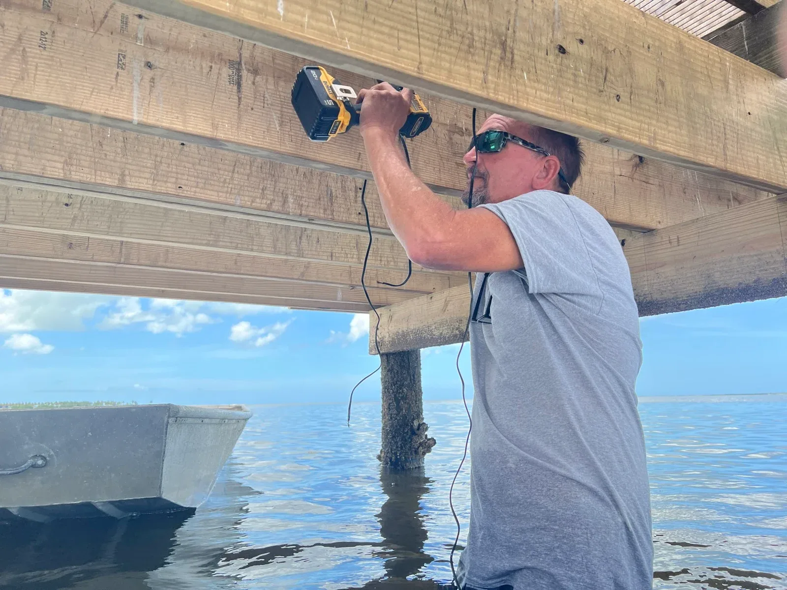 Architectural Ambiance installer drilling into a dock joist to run low-voltage lighting wire, standing in shallow water beside a barnacled piling