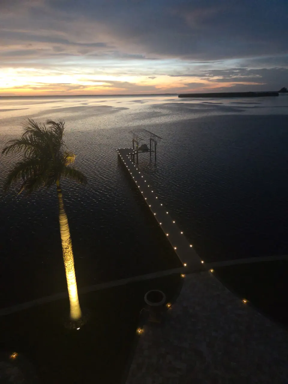 Long private dock lit with recessed deck lights running its full length at sunset, an uplit palm alongside and a boat lift at the end over open water